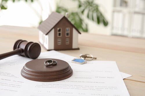 Papers underneath a gavel with two rings & a house in the background to symbolize Texas property division & divorce.